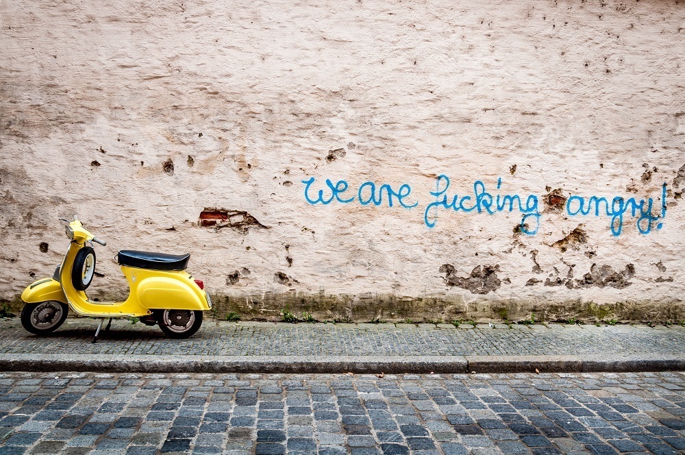 Graffiti on city wall saying We Are Fucking Angry! next to a yellow moped