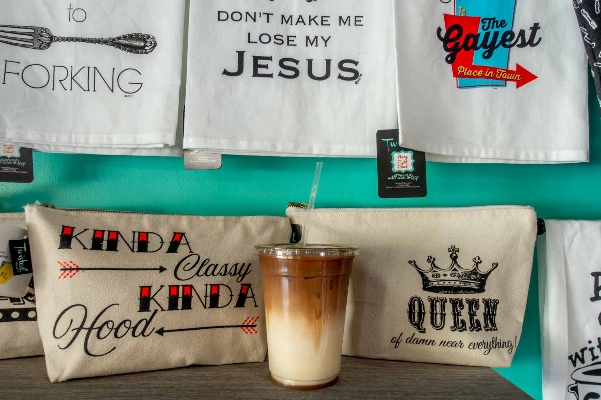 Coffee drink on shelf surrounded by bags and towels with humorous sayings. 