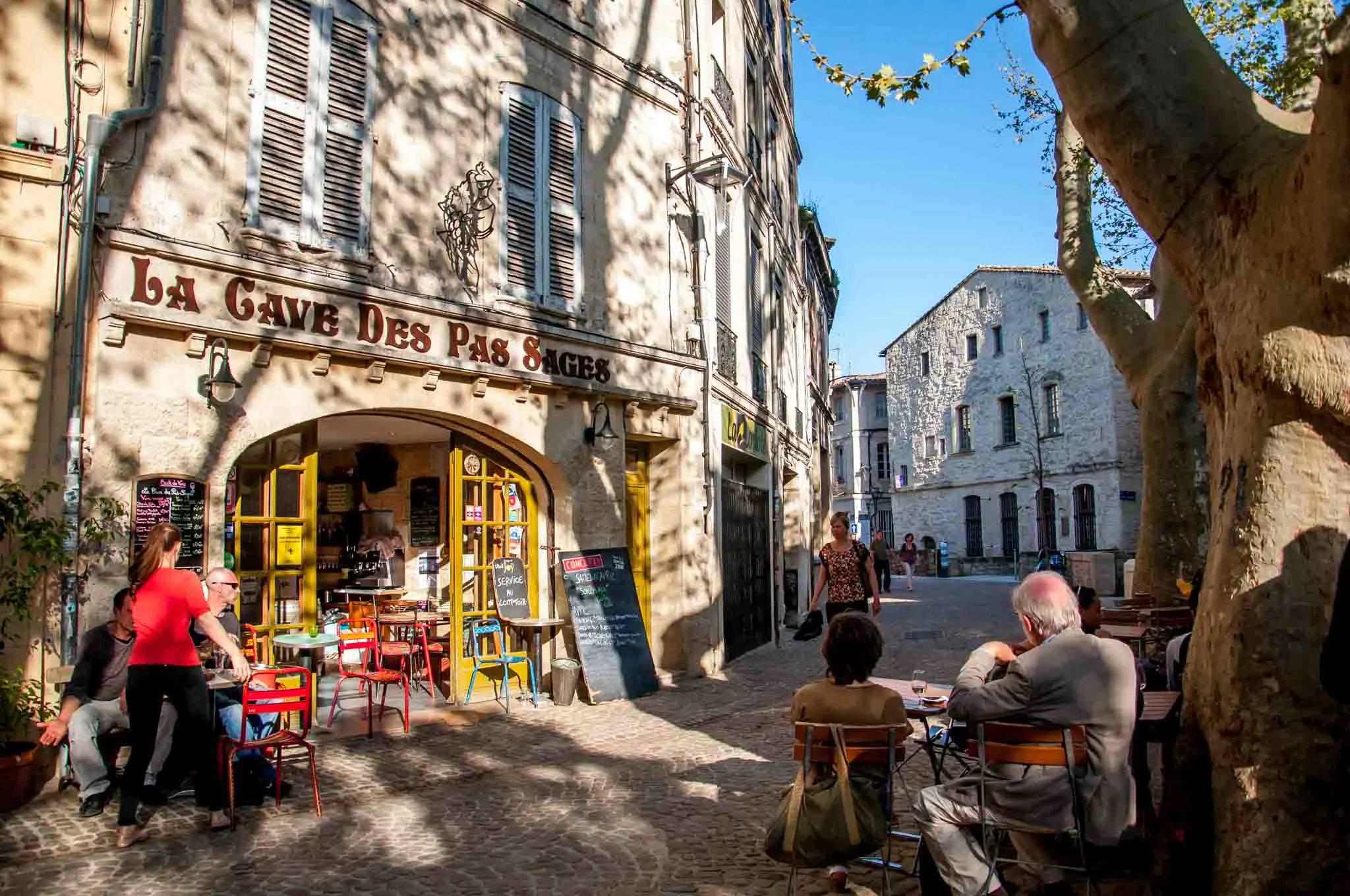 People lounging outside a cafe with open doors and a sign for "La Cave des Pas Sage." 