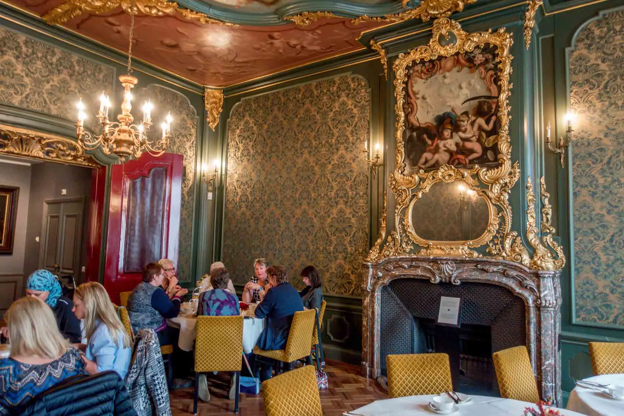 People in tea room decorated with brocade wallpaper and ornate fireplace