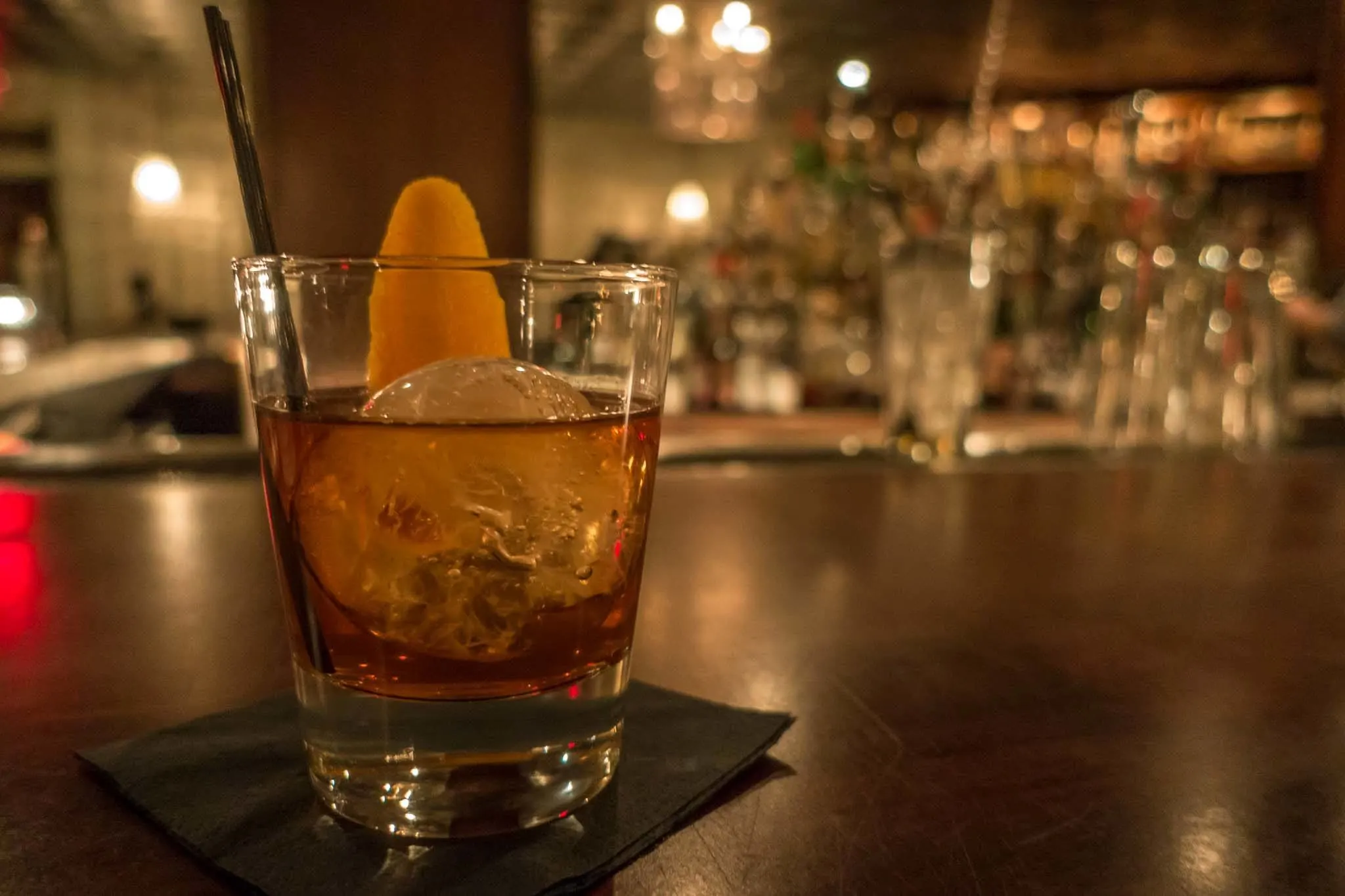 Whiskey in a glass with a large ice cube and orange garnish sitting on a wooden bar