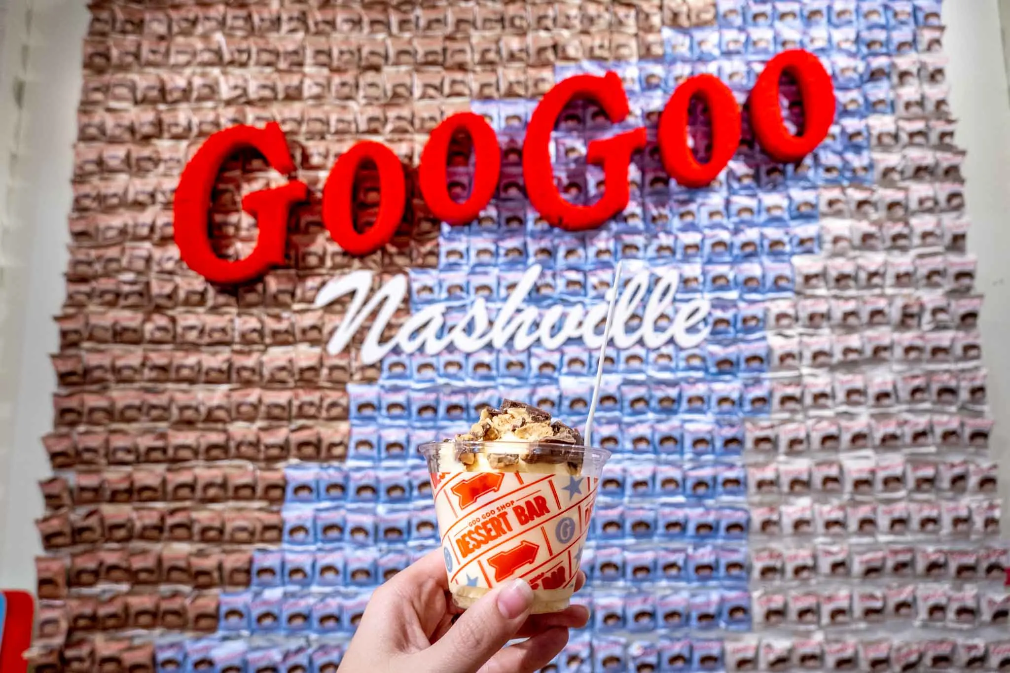 Hand holding a cup of ice cream topped with candy in front of a decorated wall with a sign: Goo Goo Nashville