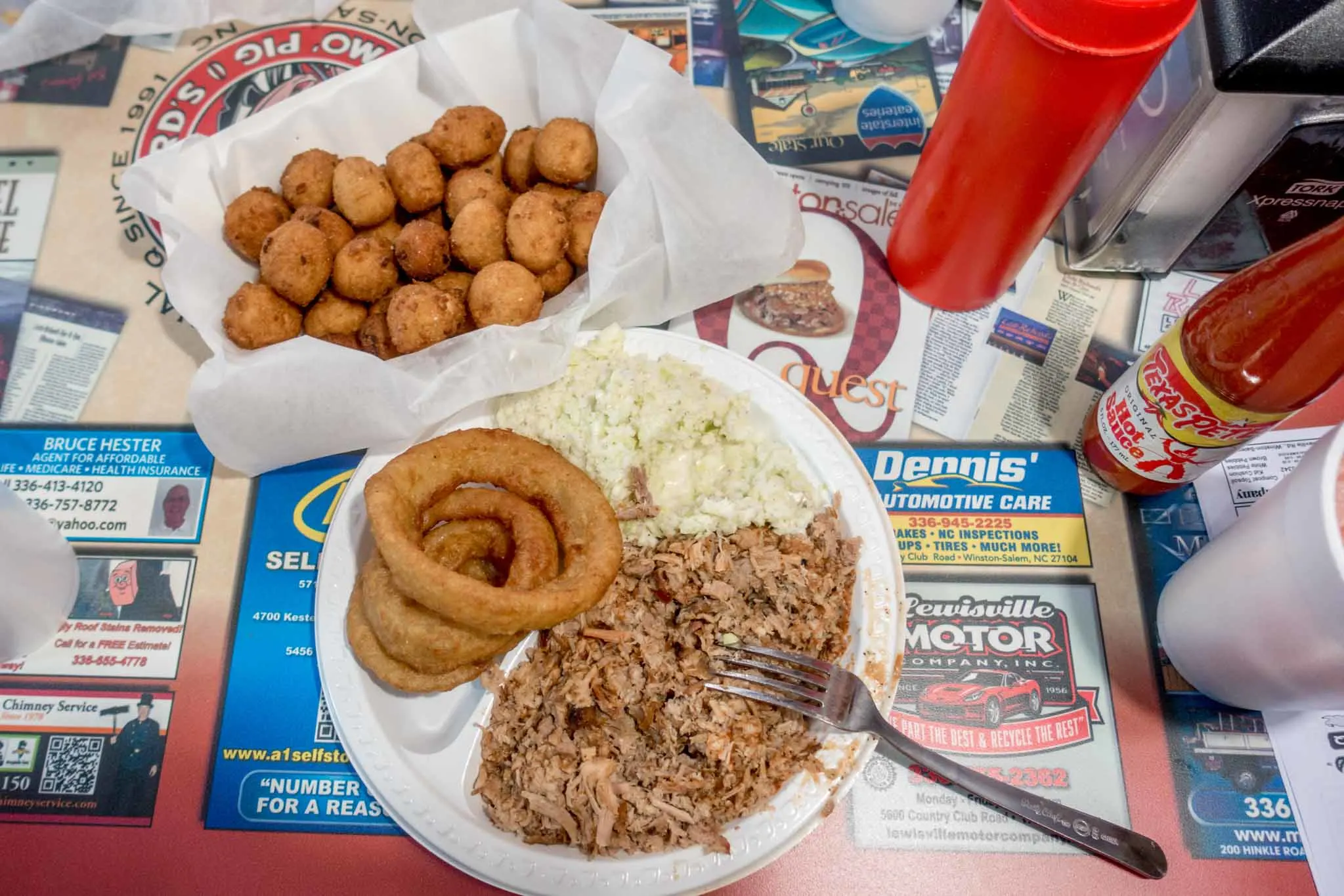 Plate of chopped barbecue, onion rings, and hush puppies on a table covered with colorful advertisements