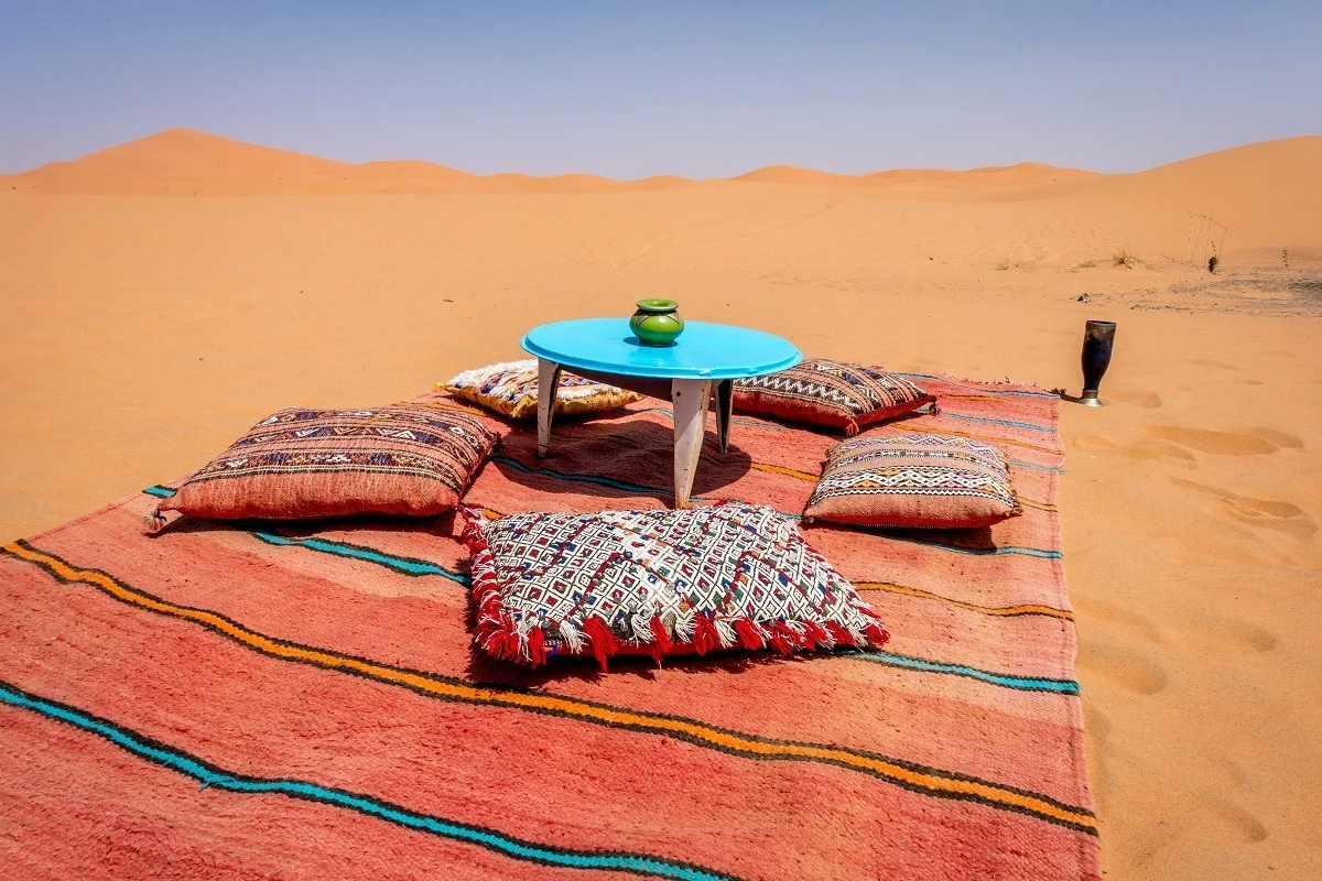 Blanket, pillows, and table set for a meal in a sand dune.