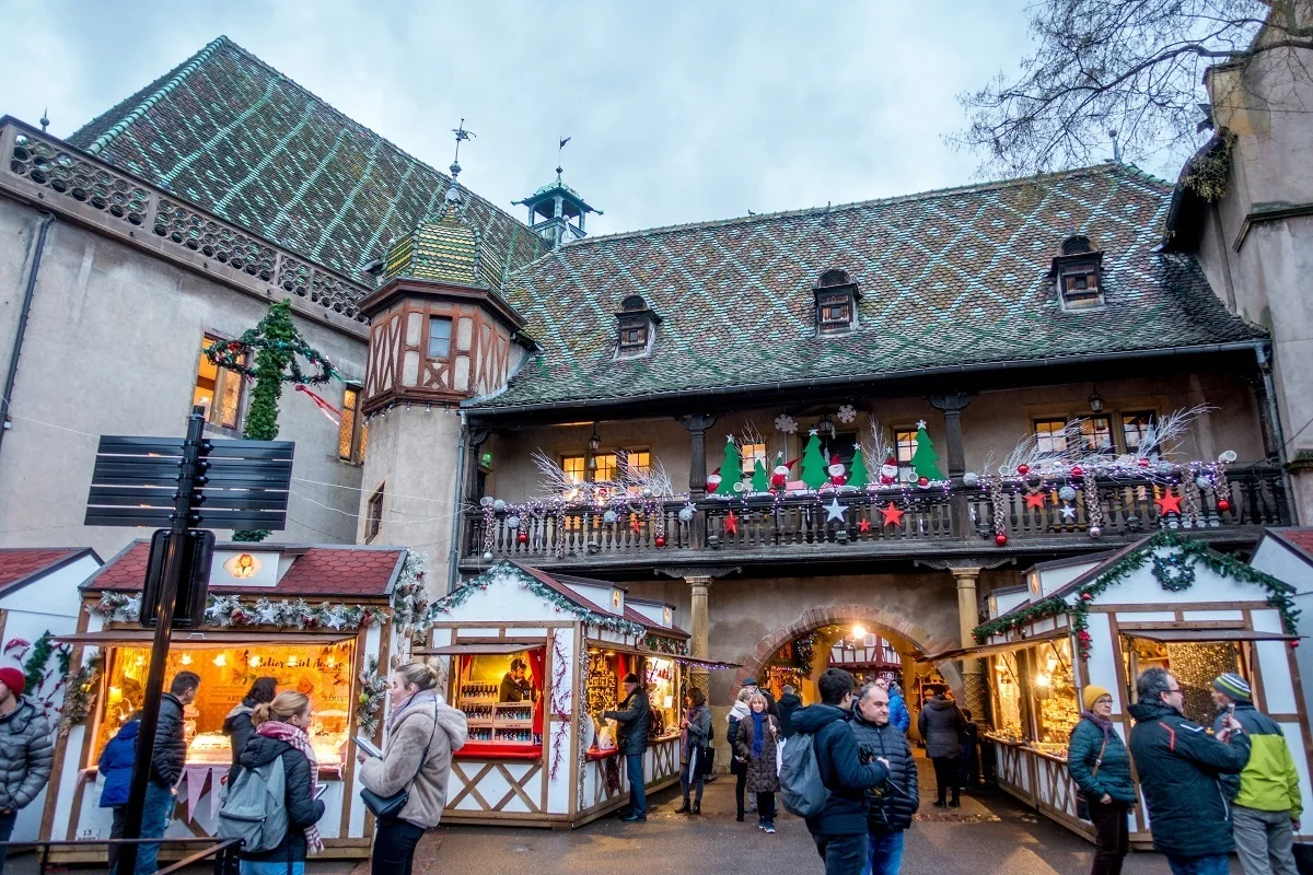 People shopping at market stalls by a building with green tile roof.