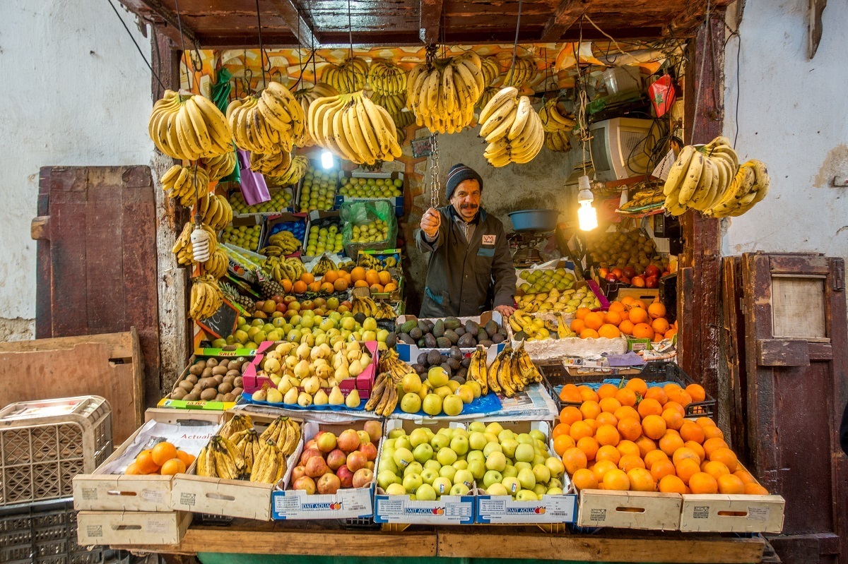 Man selling fruit at a stand filled with bananas, oranges, and more.