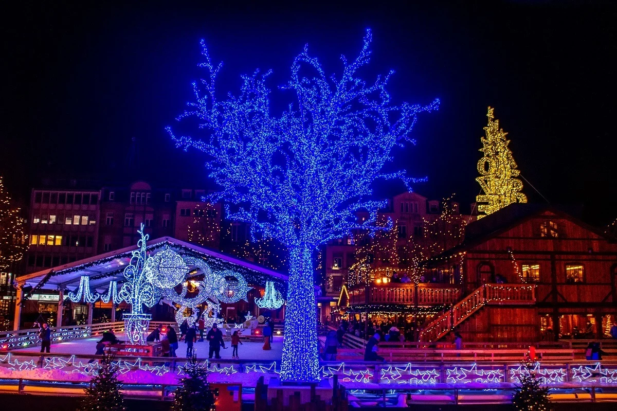 Illuminated ice skating rink, tree, and food stalls at night in Luxembourg City.