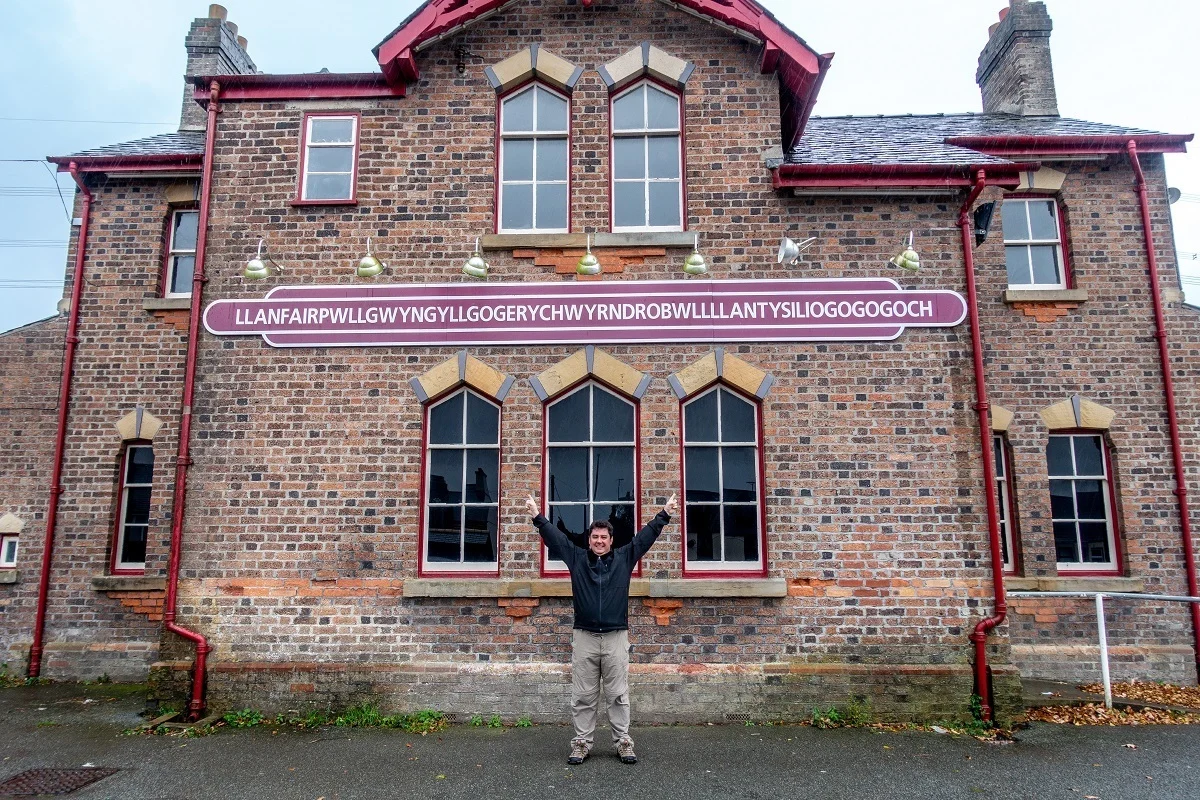 Person in front of a brick building with a sign for Llanfairpwllgwyngyllgogerychwyrndrobwllllantysiliogogogoch 