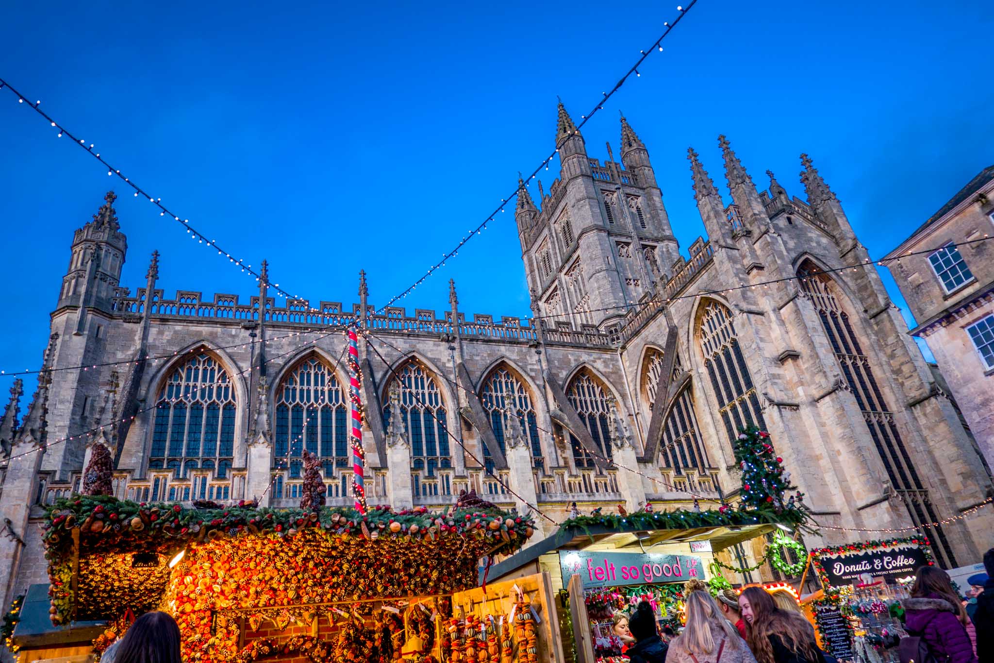 Stalls of the Bath Christmas market lit up at sunset in front of the large stone Bath Abbey