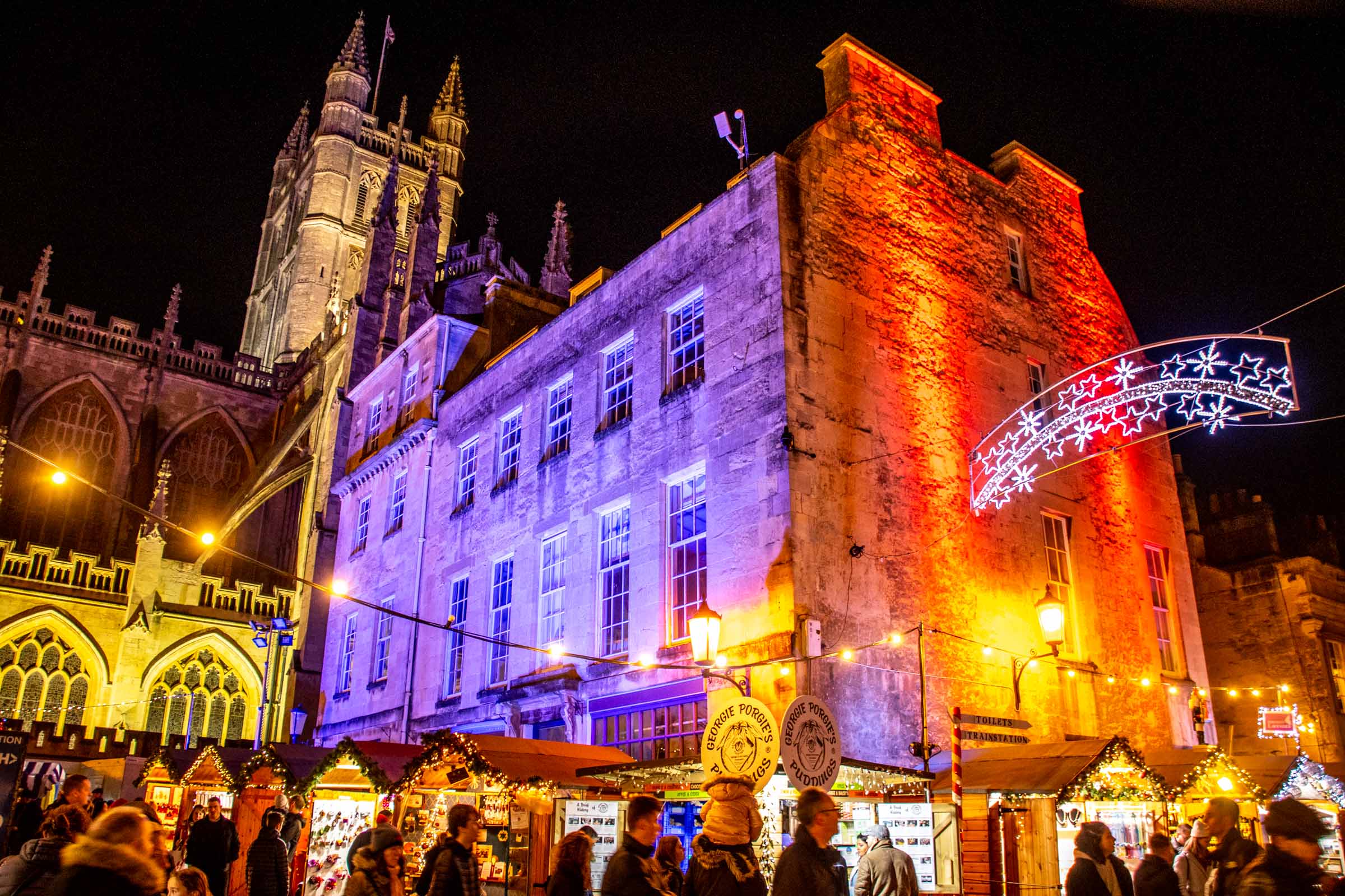 People shopping at night by Bath Abbey, lit up in purple and red lights