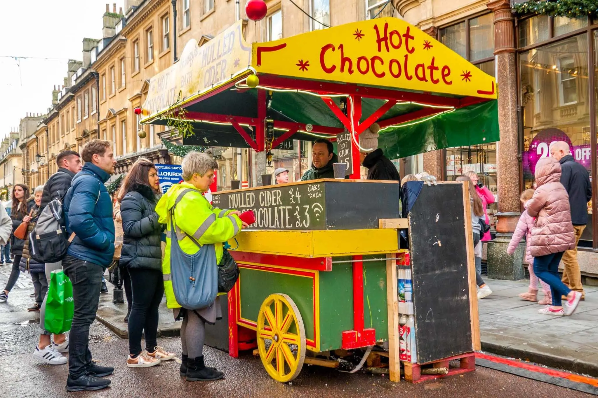 People standing at yellow and green hot chocolate cart in the street 