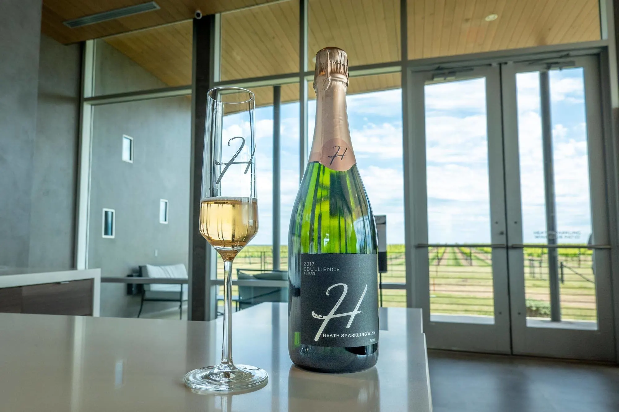 Glass and bottle of sparkling wine on a counter with a view of grapevines outside