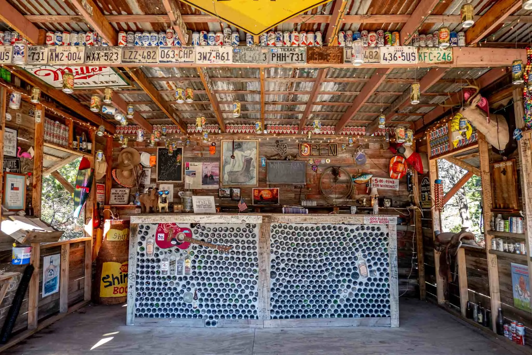 A bar made of glass bottles in a room covered with signs and beer cans.