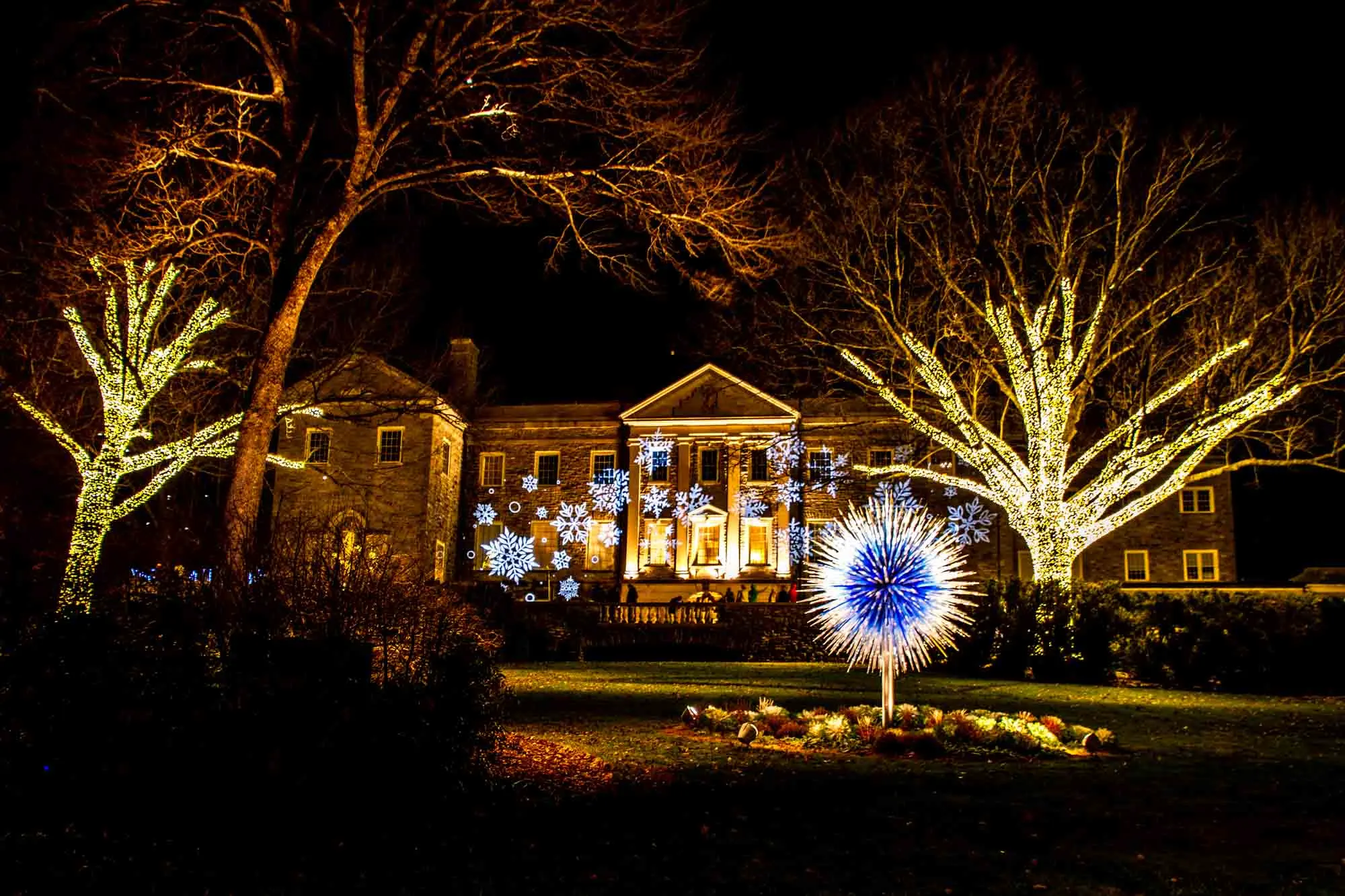 Snowflake lights projected on a mansion facade beside lit up trees and an illuminated blue and white sculpture