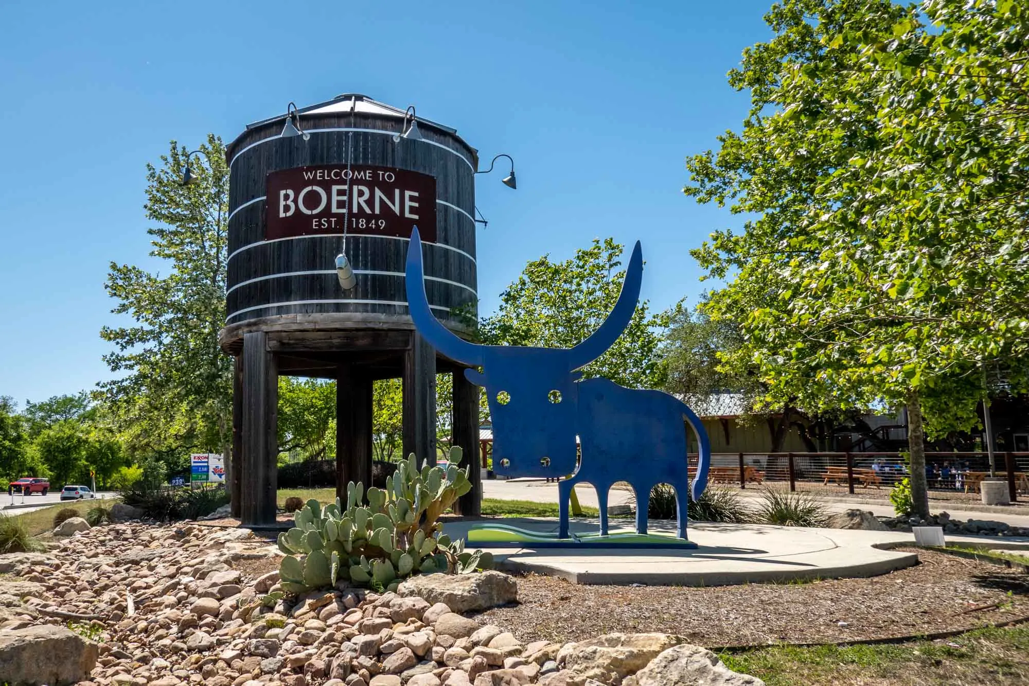 Sculpture of a blue bull with a cartooonish appearance next to a water tower with the sign "Welcome to Boerne, Est. 1849."