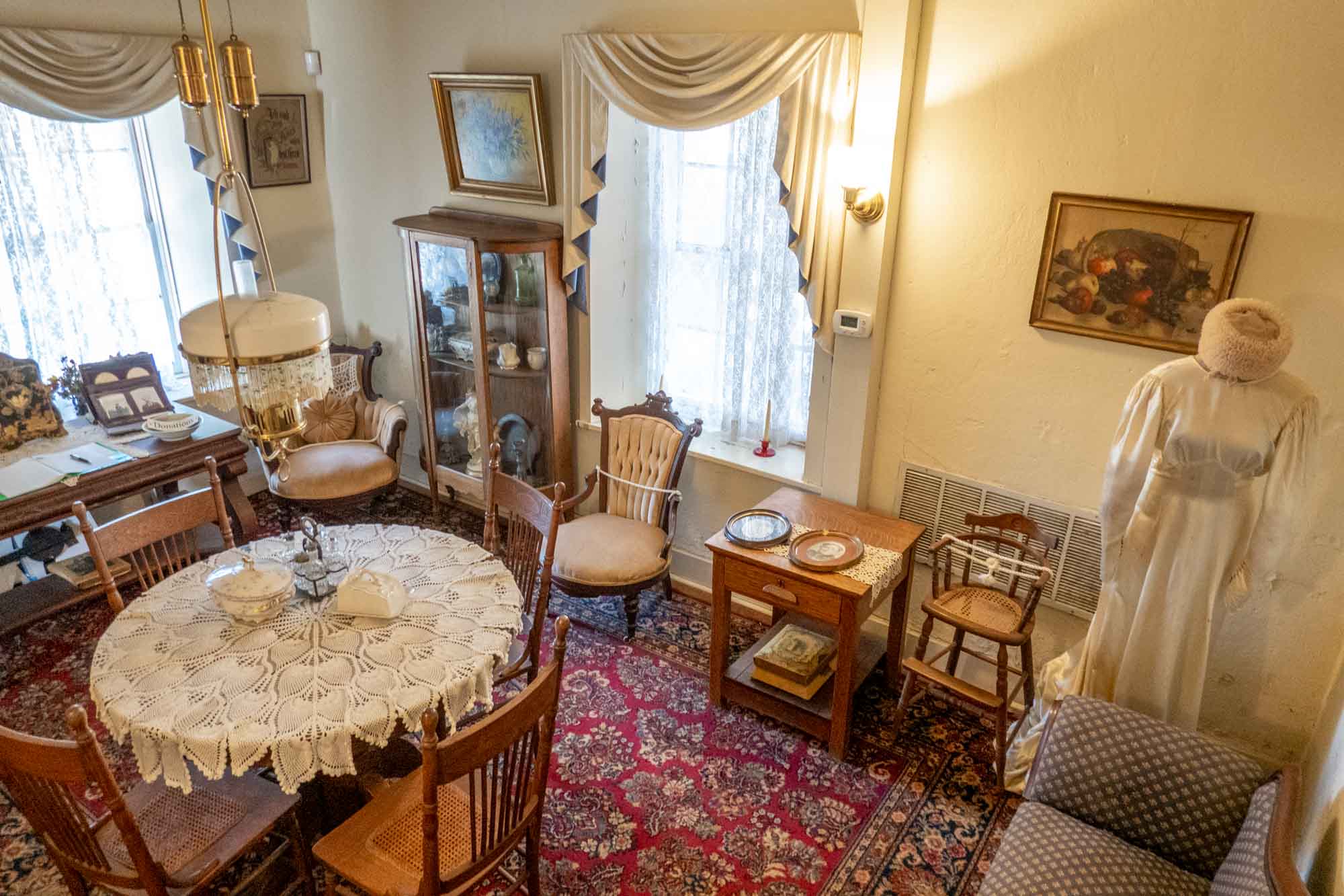 Room with antique table, chairs, dishes, and an old-fashioned white dress on display