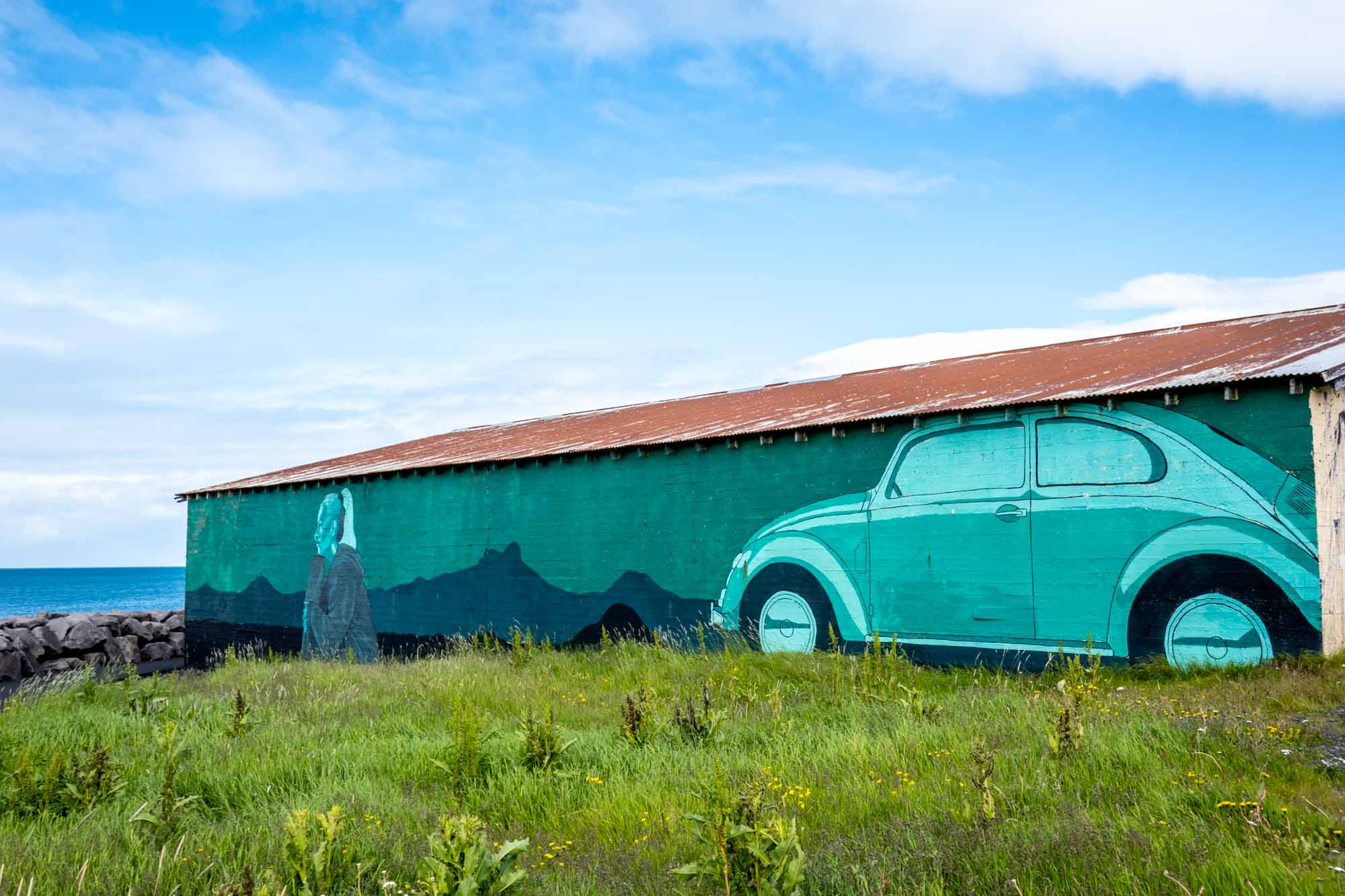 Green mural on side of building of a woman sitting in front of a VW Beatle looking out at the ocean