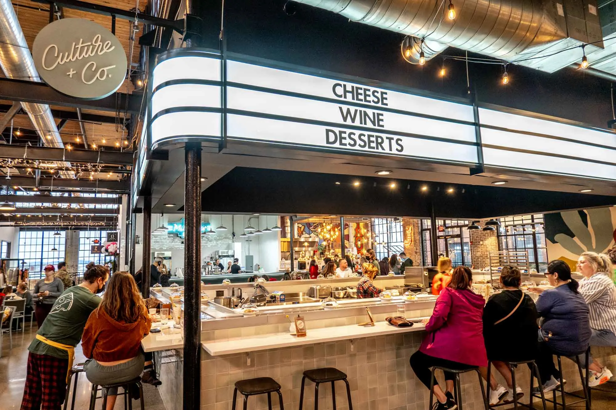 People sitting at a counter under a sign for "Culture + Co.: Cheese, Wine, Desserts"
