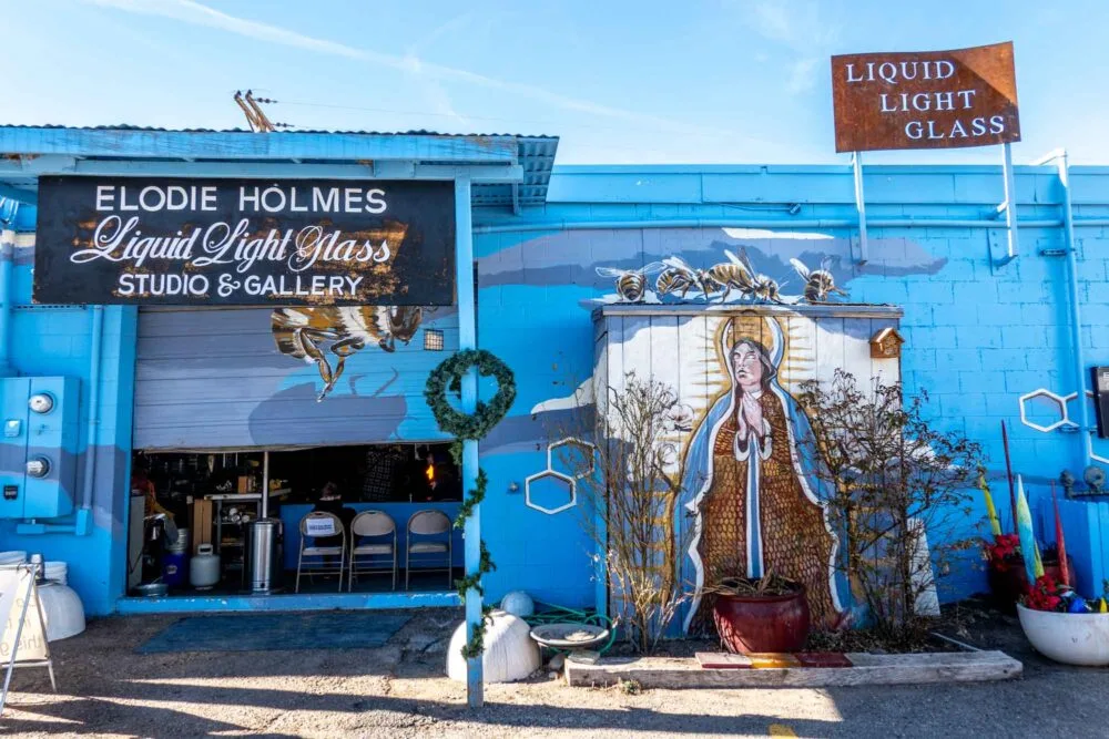 Exterior of a blue building with a mural of a woman and bees and a sign: "Elodie Holmes Liquid Light Glass Studio & Gallery."