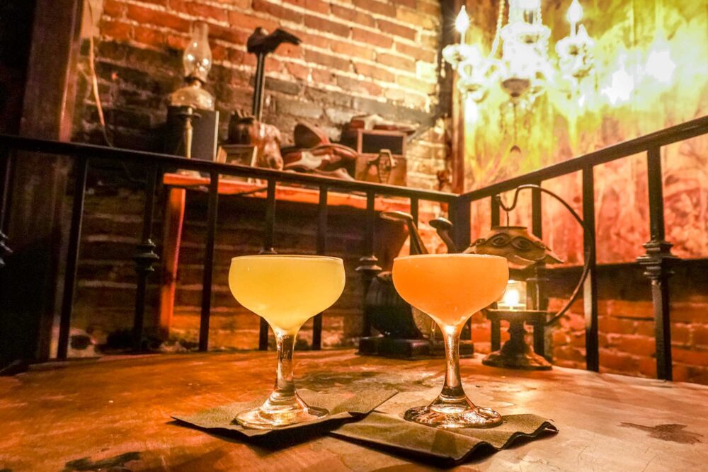 Two cocktails on a table in a dimly-lit speakeasy