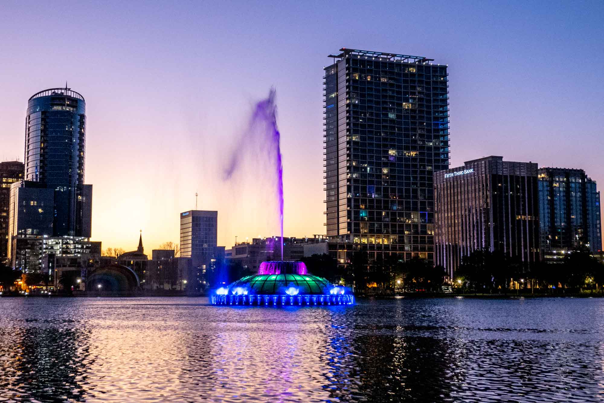 Fountain lit up in bright colors in the middle of a lake with skyscrapers in the background.