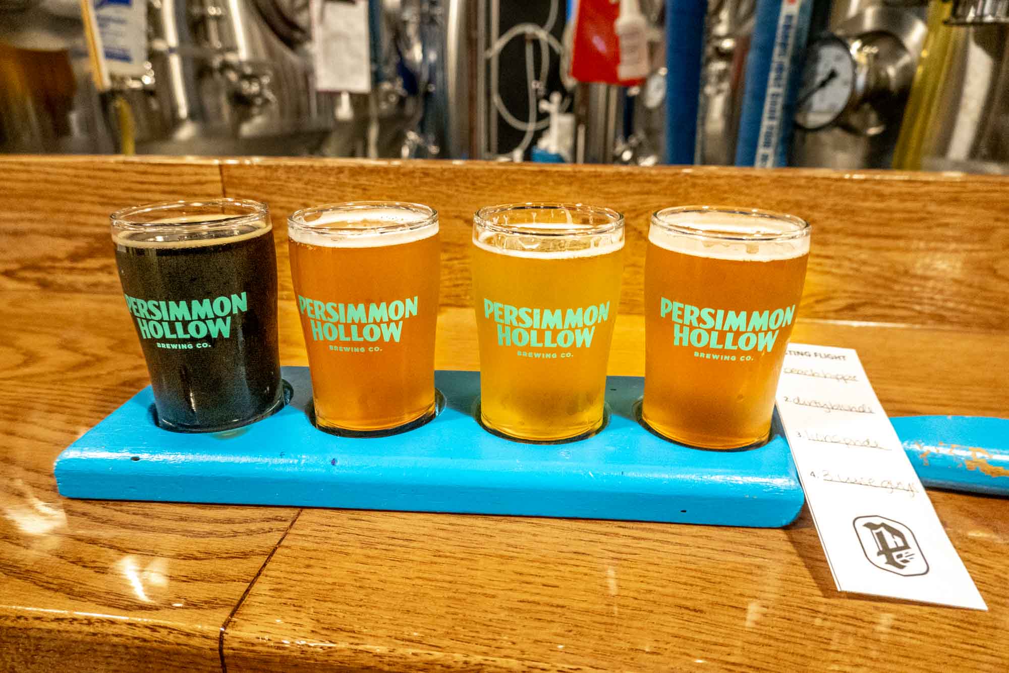 Flight of four beer glasses labeled "Persimmon Hollow Brewing Co." on a wooden bar.