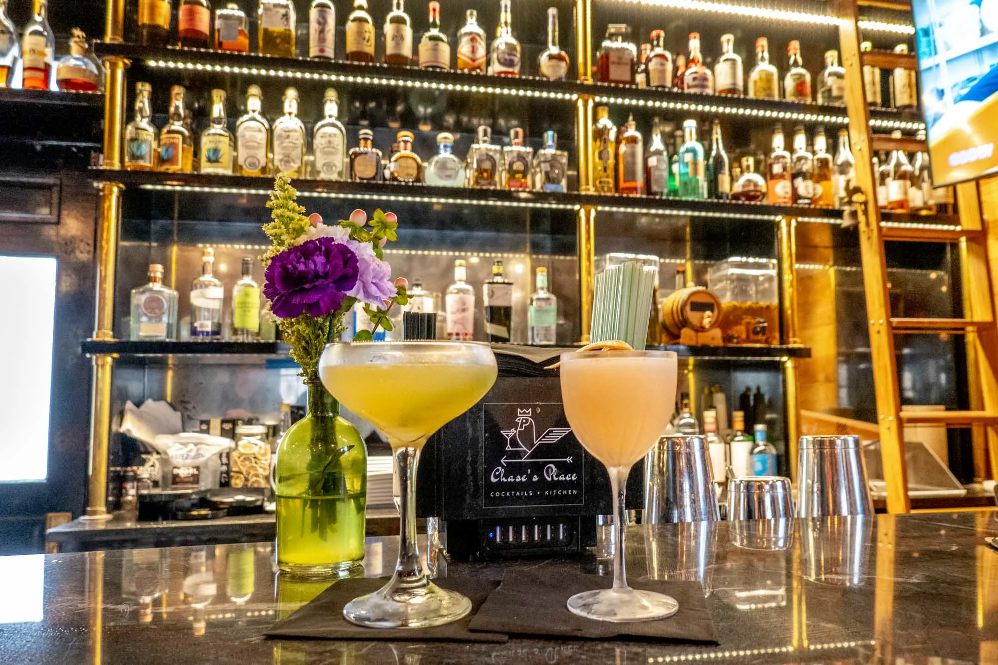 Two cocktails on a bar in front of shelves stocked with alcohol bottles