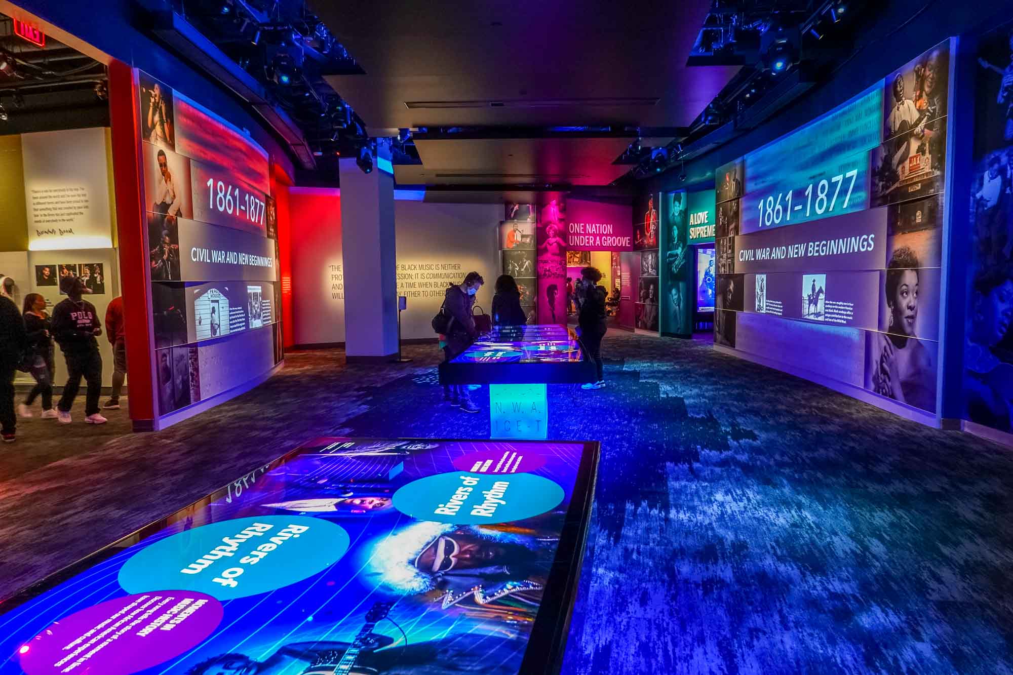 People standing at illuminated tables in a dark room filled with colorful, lit up exhibits.