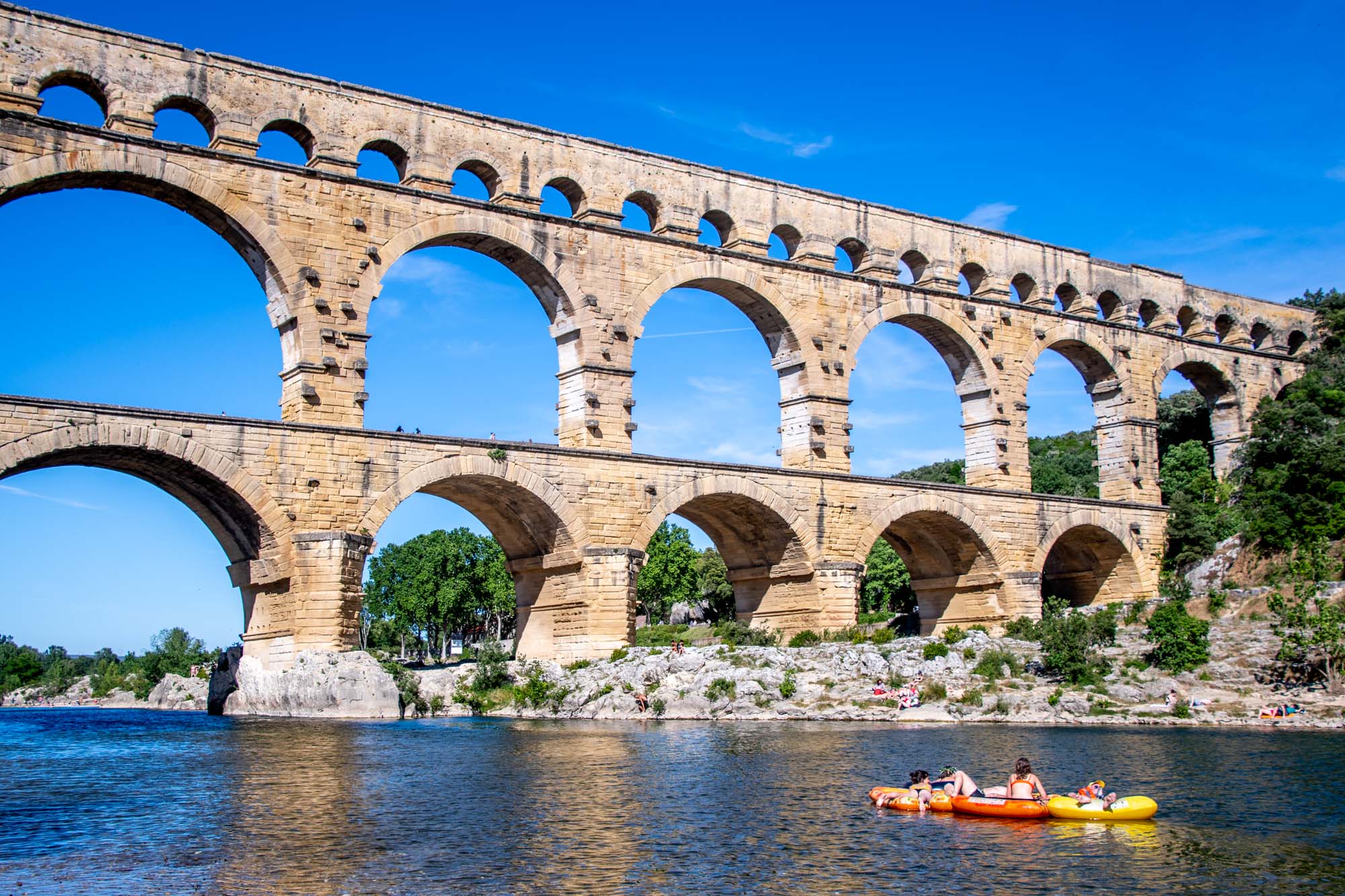 Three-level ancient Roman aqueduct towering over a river with people in a raft