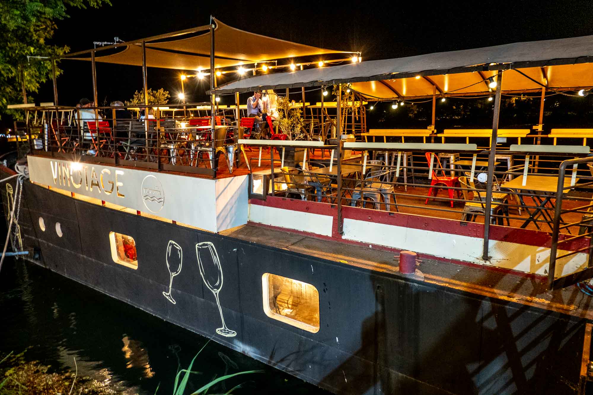 Wine bar on a black barge lit up at night with a sign for "Vinotage."