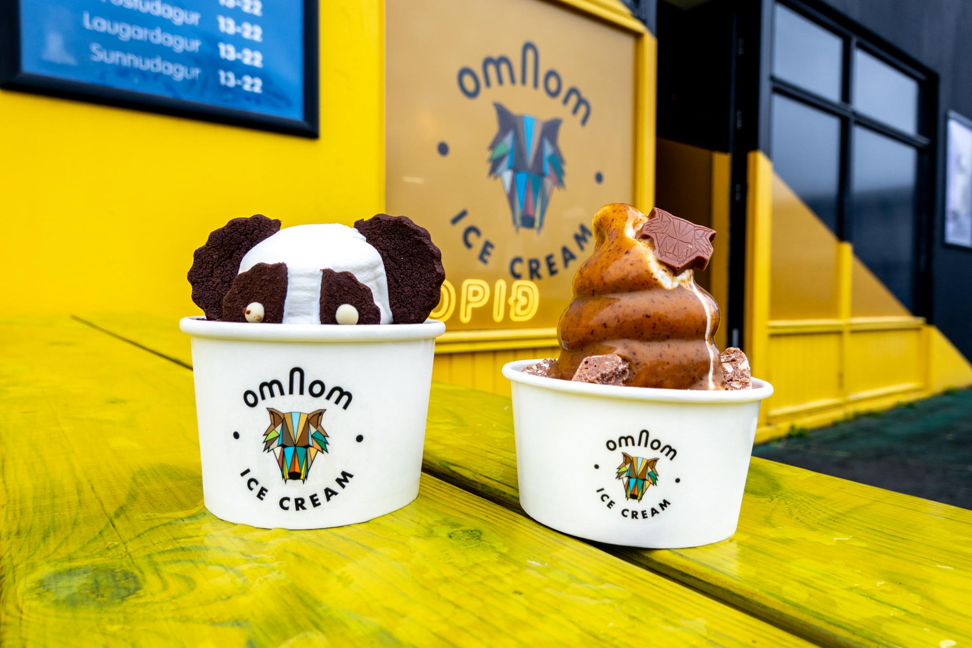 Ice cream in cups on a yellow picnic table in front of a sign for Omnom ice cream.