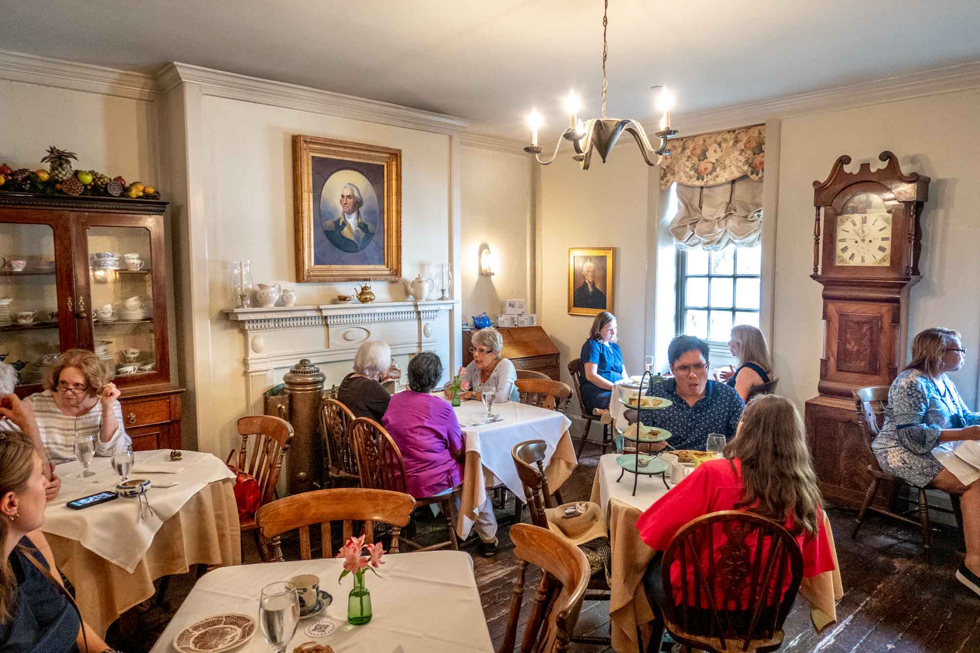 People eating at tables in a Colonial-style dining room with a portrait of George Washington on the wall.