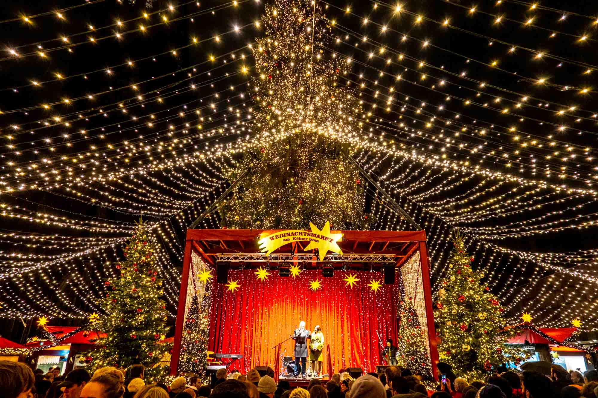 Singers on a stage under a canopy of Christmas lights