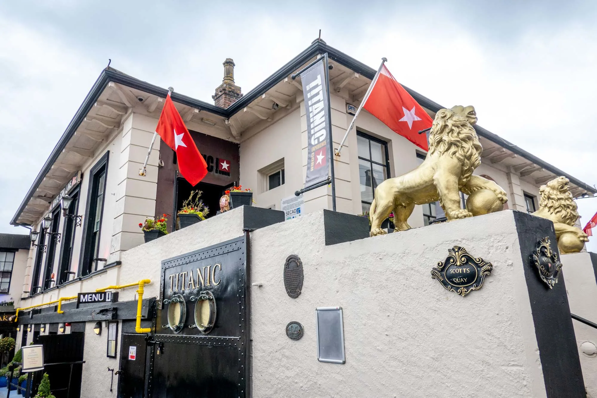 Gray concrete building with two golden lion statues and red flags with a white star by a sign for "Titanic Experience"