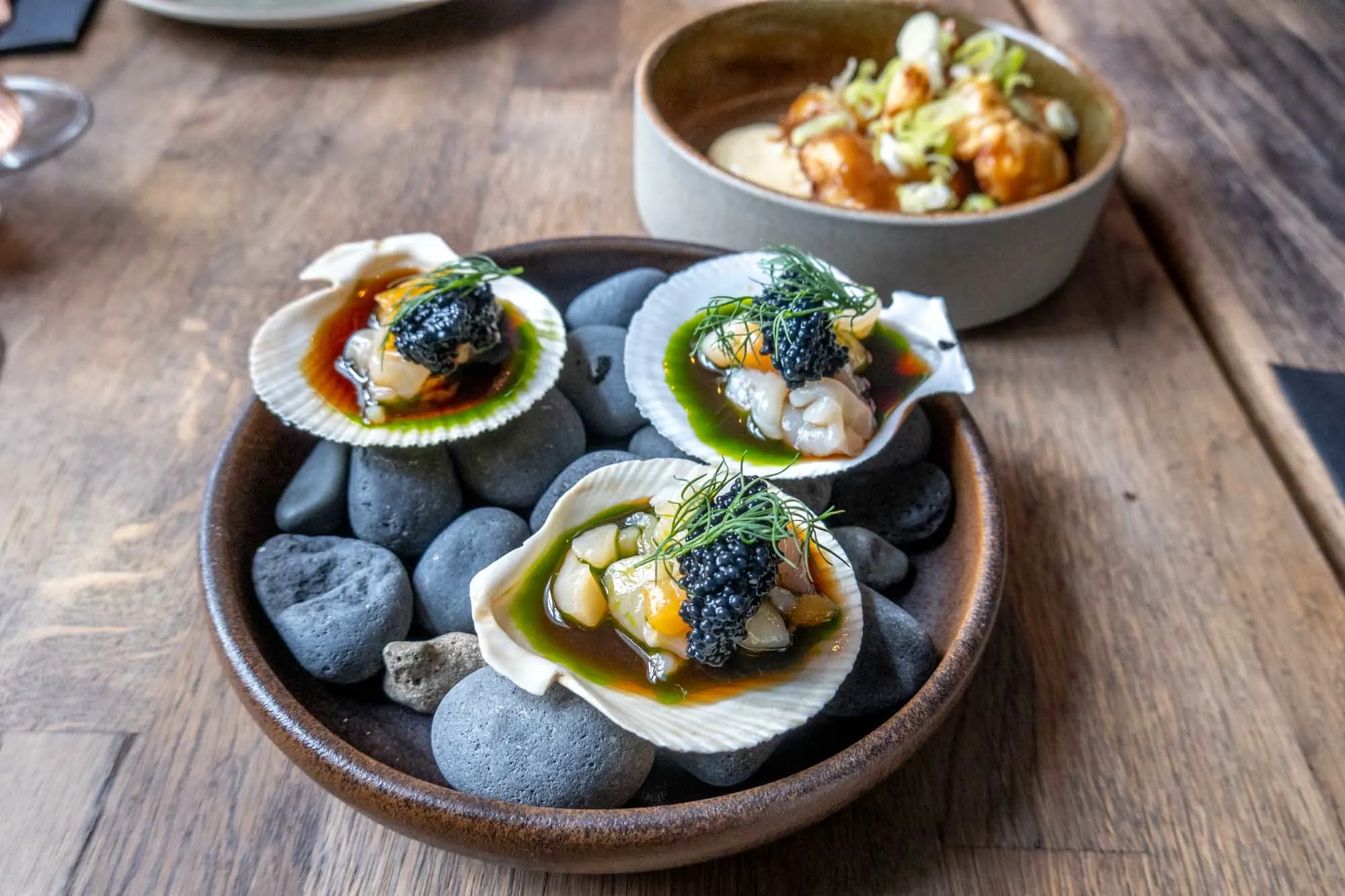 Scallops and caviar served in a scallop shell on a bed of black lava rocks in a clay bowl