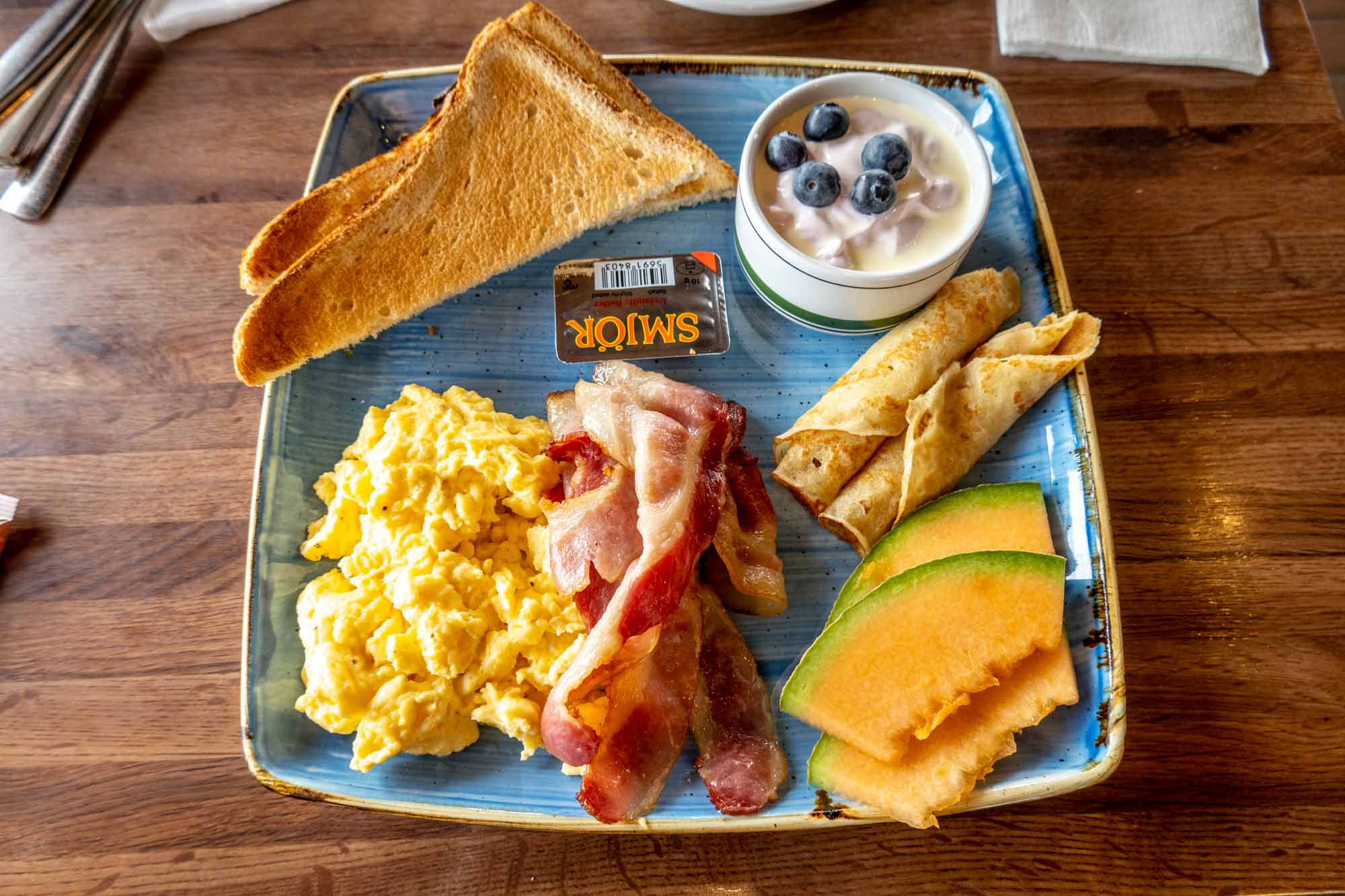 A blue plate filled with eggs, toast, bacon, crepes, yogurt and melon