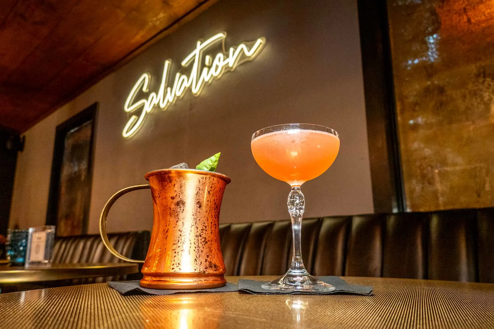 Two cocktails on a table in front of a sign for "Salvation."