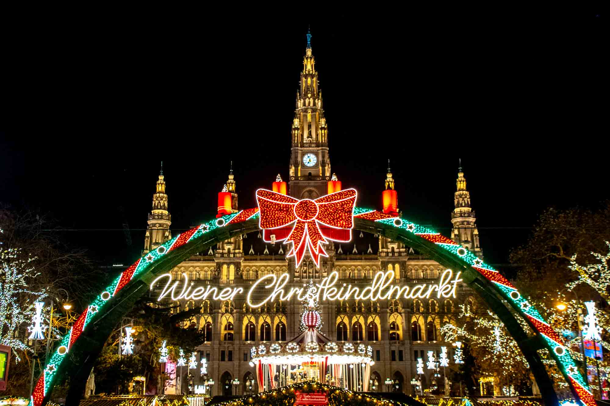 Ornate building lit up at night behind an arch of Christmas lights and sign for "Wiener Christkindlmarkt" (Vienna Christmas market).