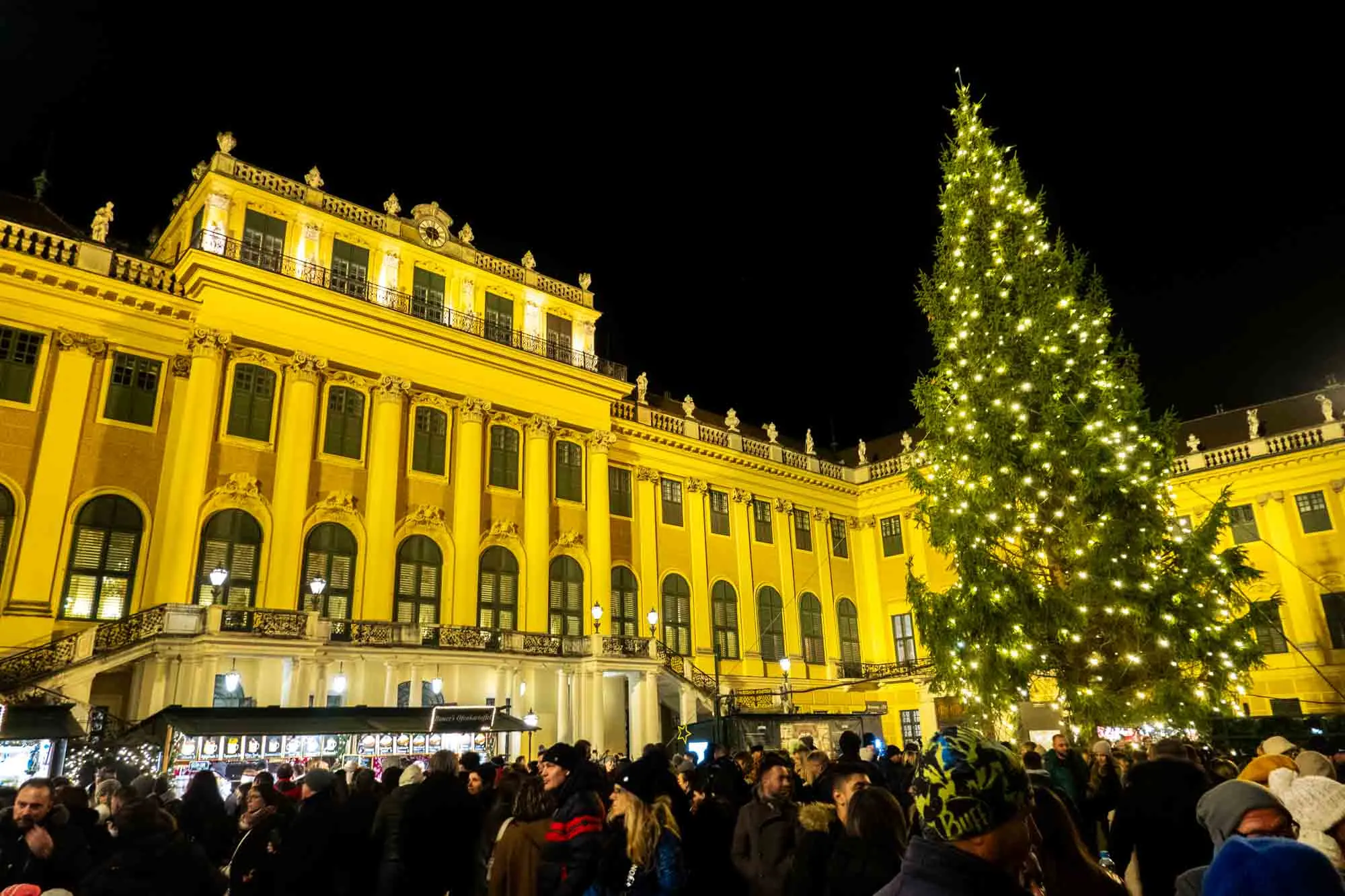 People milling around a Christmas tree in front of a large yellow building.
