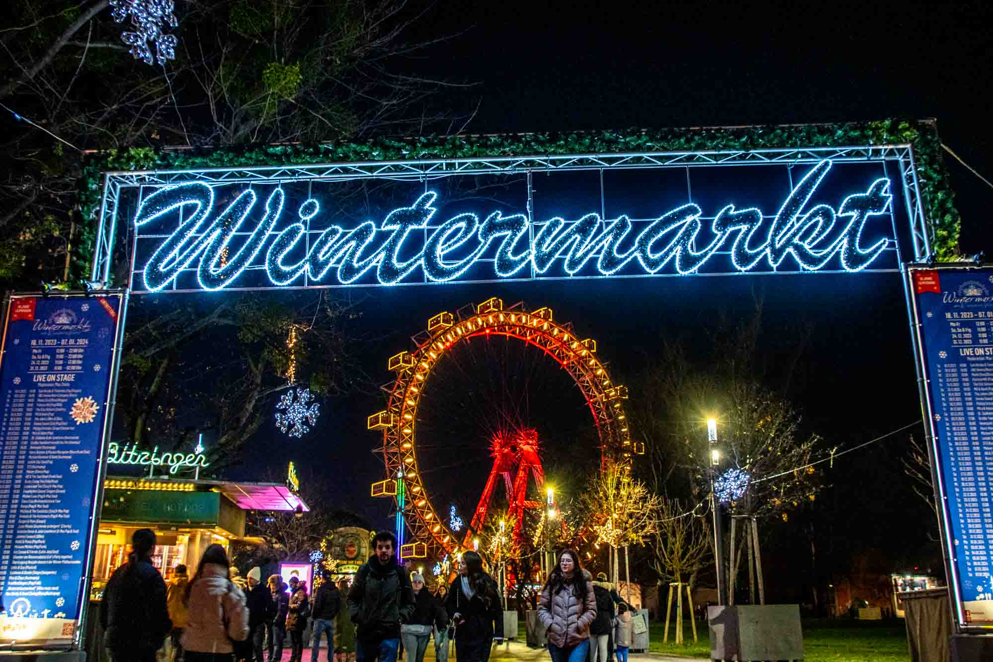 People walking by an illuminated Ferris wheel framed by an illuminated sign for "Wintermarkt" (Winter Market).