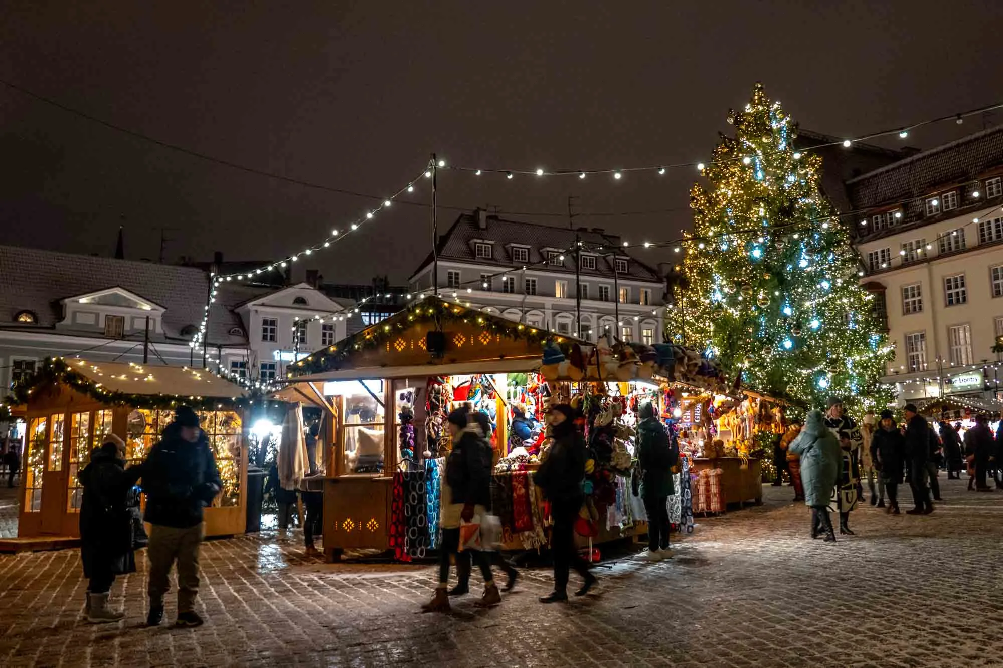 Shoppers passing wooden Christmas market stalls at night with a Christmas tree in the background.